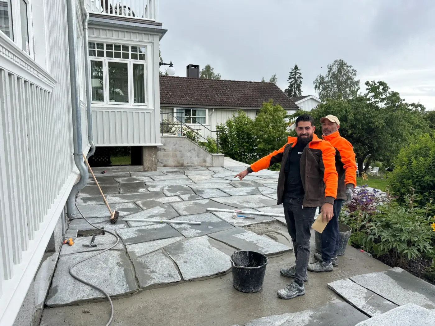 Grå naturstein-terrasse under anlegging med fuktig plenområde og omgivende heller.
