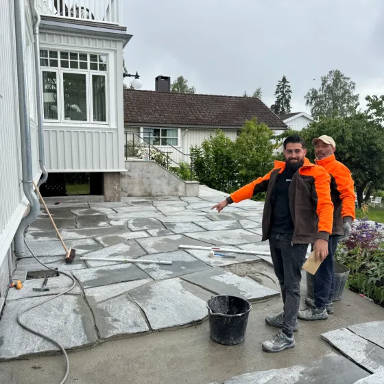 Grå naturstein-terrasse under anlegging med fuktig plenområde og omgivende heller.
