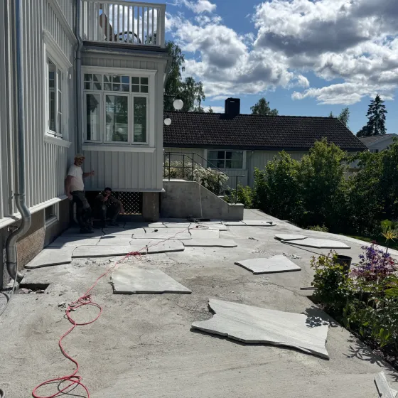 Terrasse under anlegg med grå heller og kantstein, omgitt av hekker og blomster.