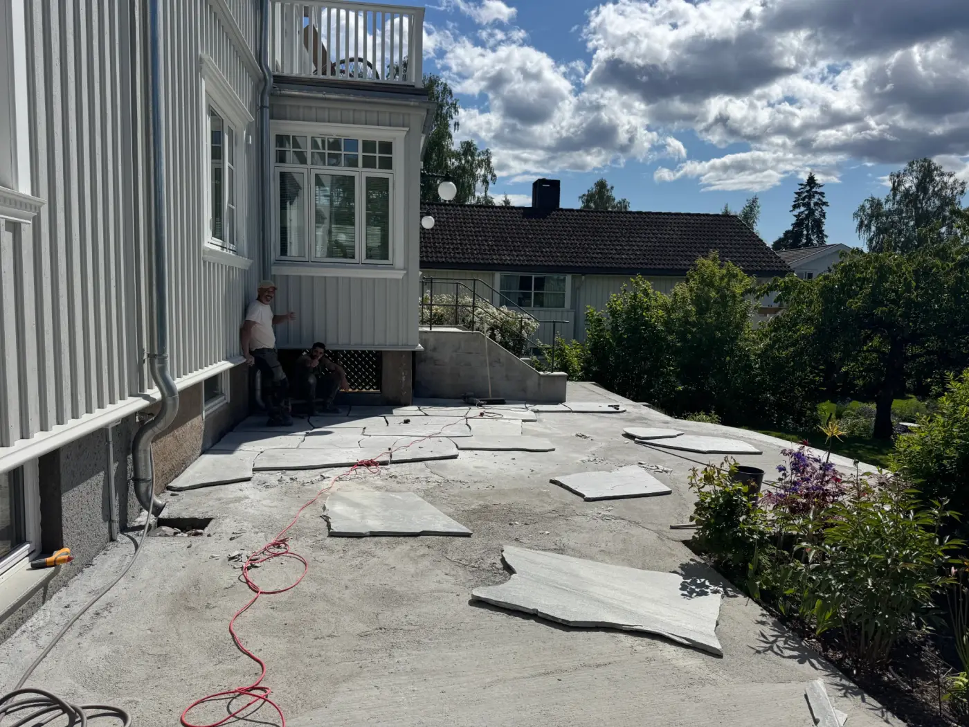 Terrasse under anlegg med grå heller og kantstein, omgitt av hekker og blomster.
