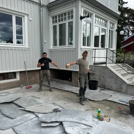 Anlegging av skifertegning på terrasse foran hvitt hus under konstruksjon.