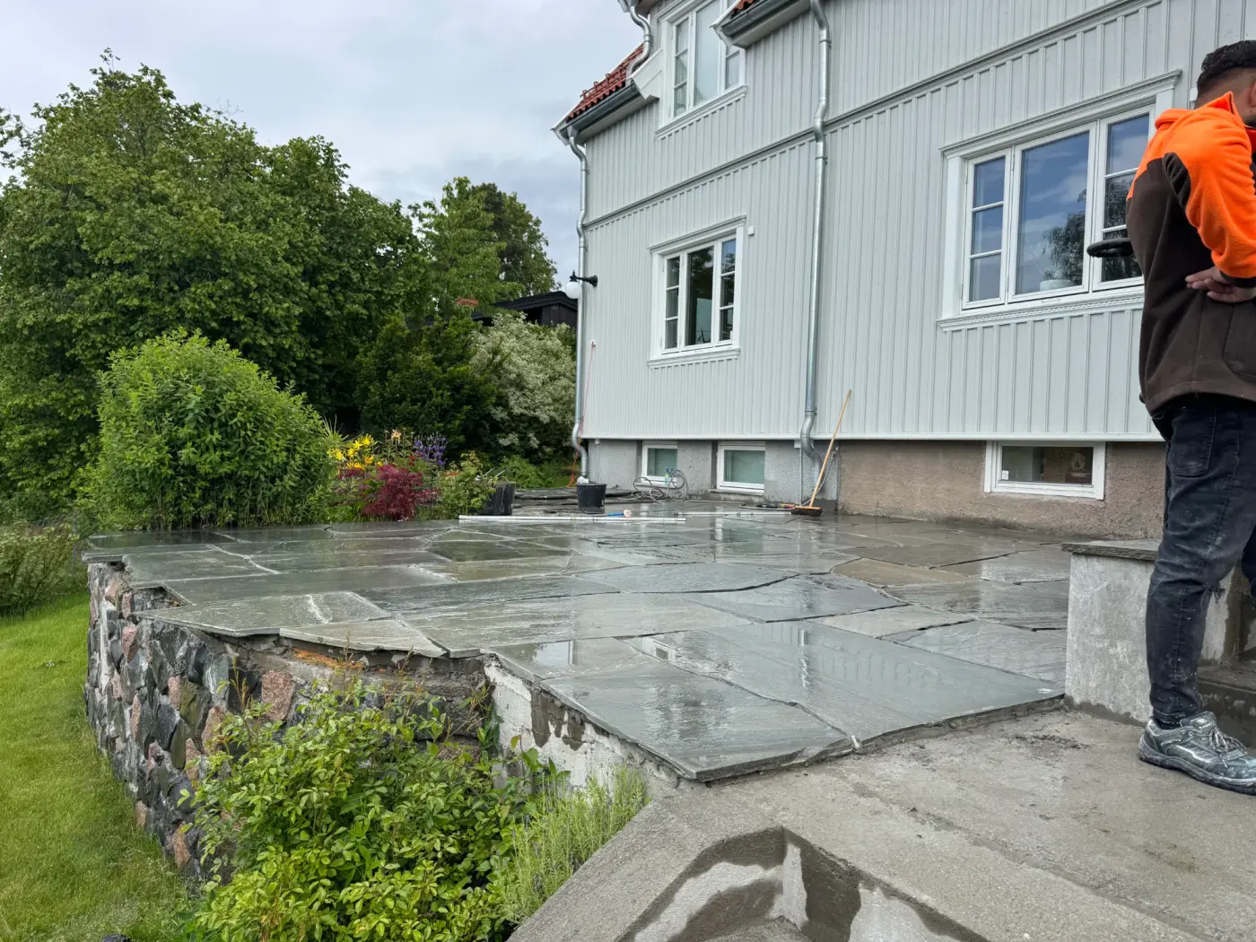 Skiferplatting på terrasse ved hus med steinmur og grønn hage.