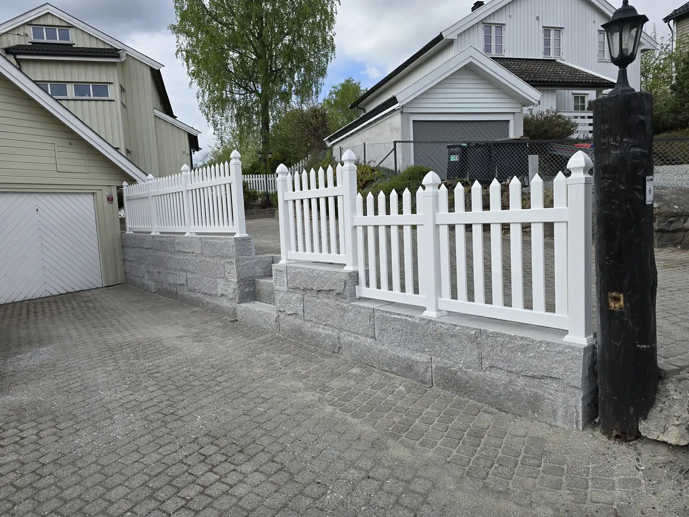Granittblokkmur med stakittgjerde over gårdsplass i smågatestein