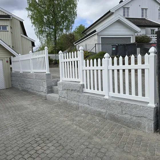 Granittblokkmur med stakittgjerde over gårdsplass i smågatestein