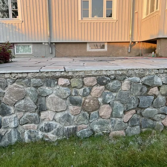 Mur av rund bruddstein som bærer en uteplass med store heller