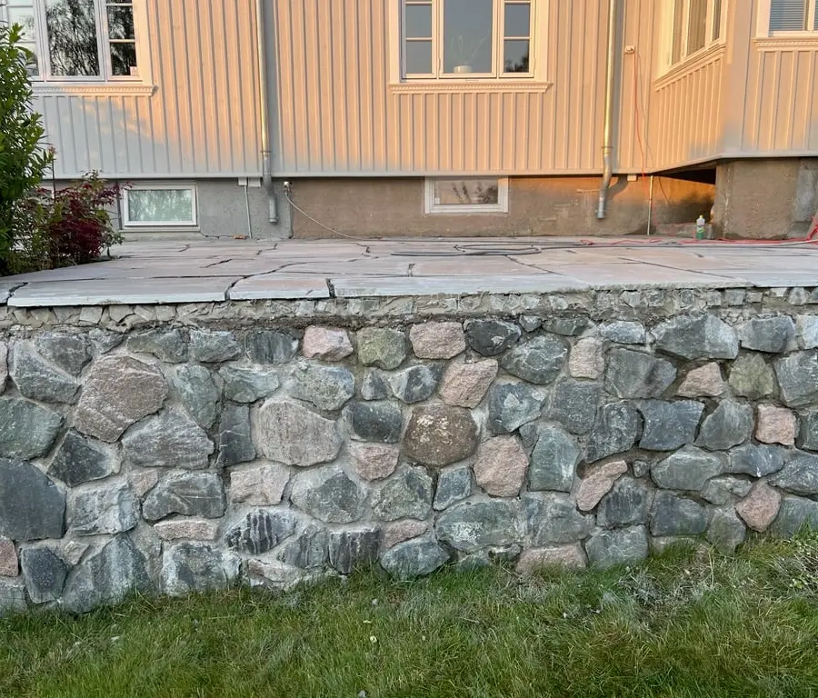 Mur av rund bruddstein som bærer en uteplass med store heller