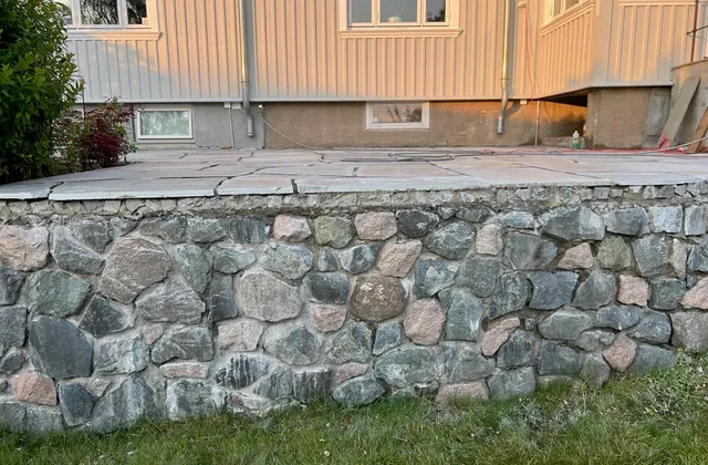 Mur av rund bruddstein som bærer en uteplass med store heller
