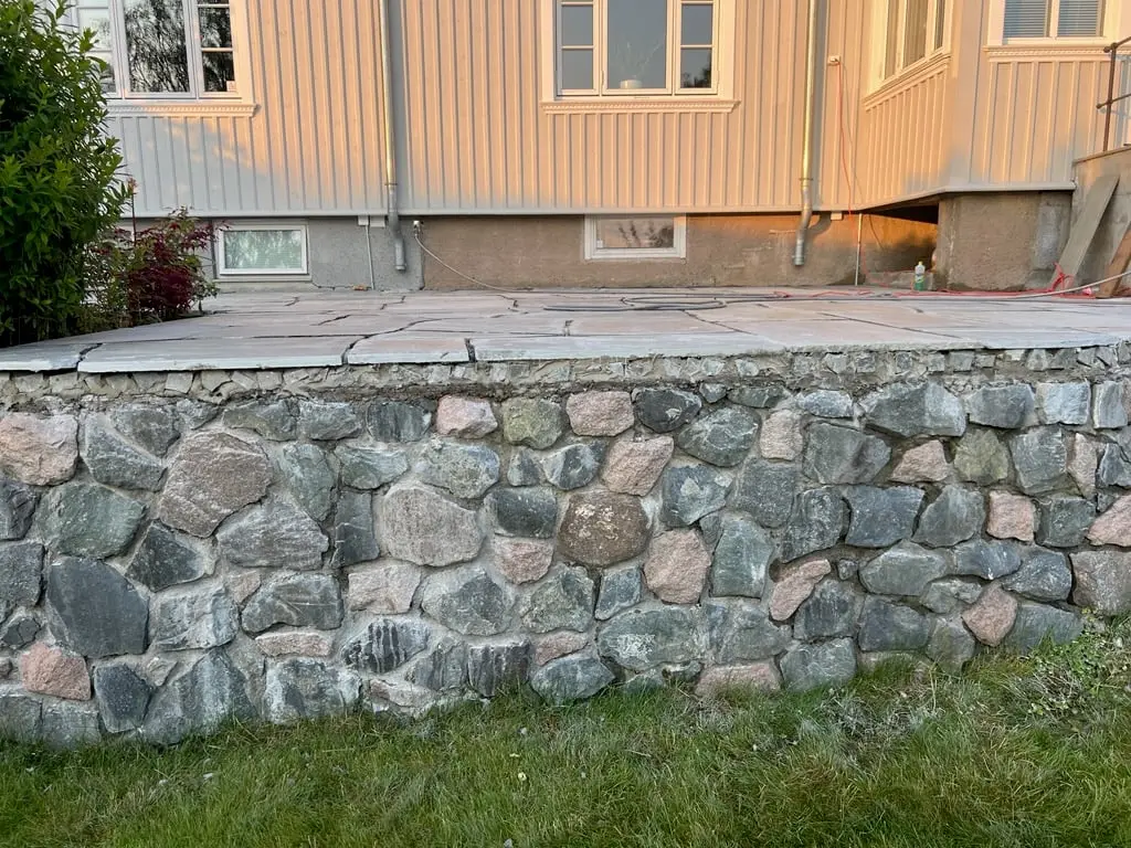 Mur av rund bruddstein som bærer en uteplass med store heller