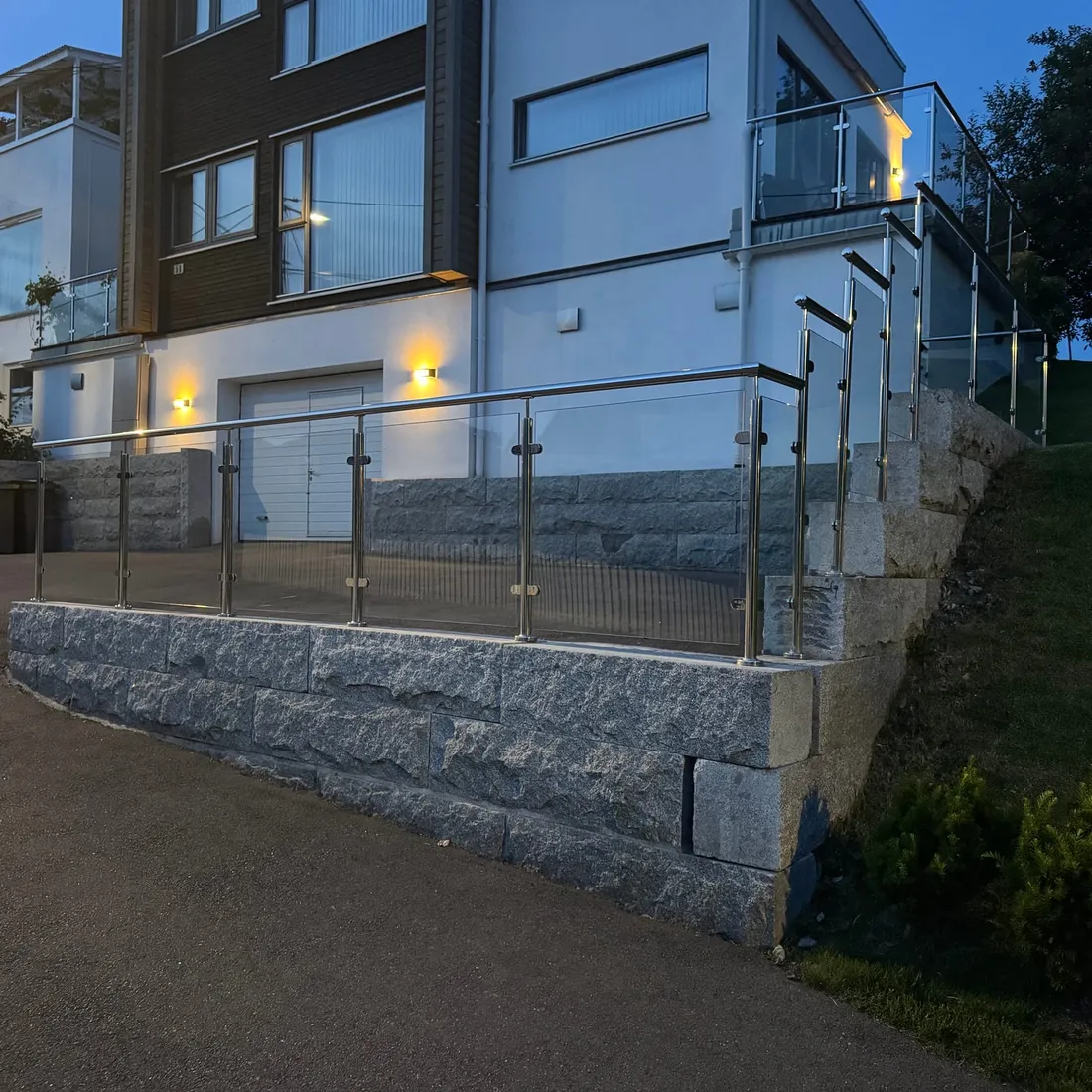 Granittblokkmur med glassrekkverk langs oppkjørsel, opplyst hus i skumringen